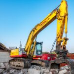 A large yellow excavator working amidst rubble at a construction site under a clear blue sky.